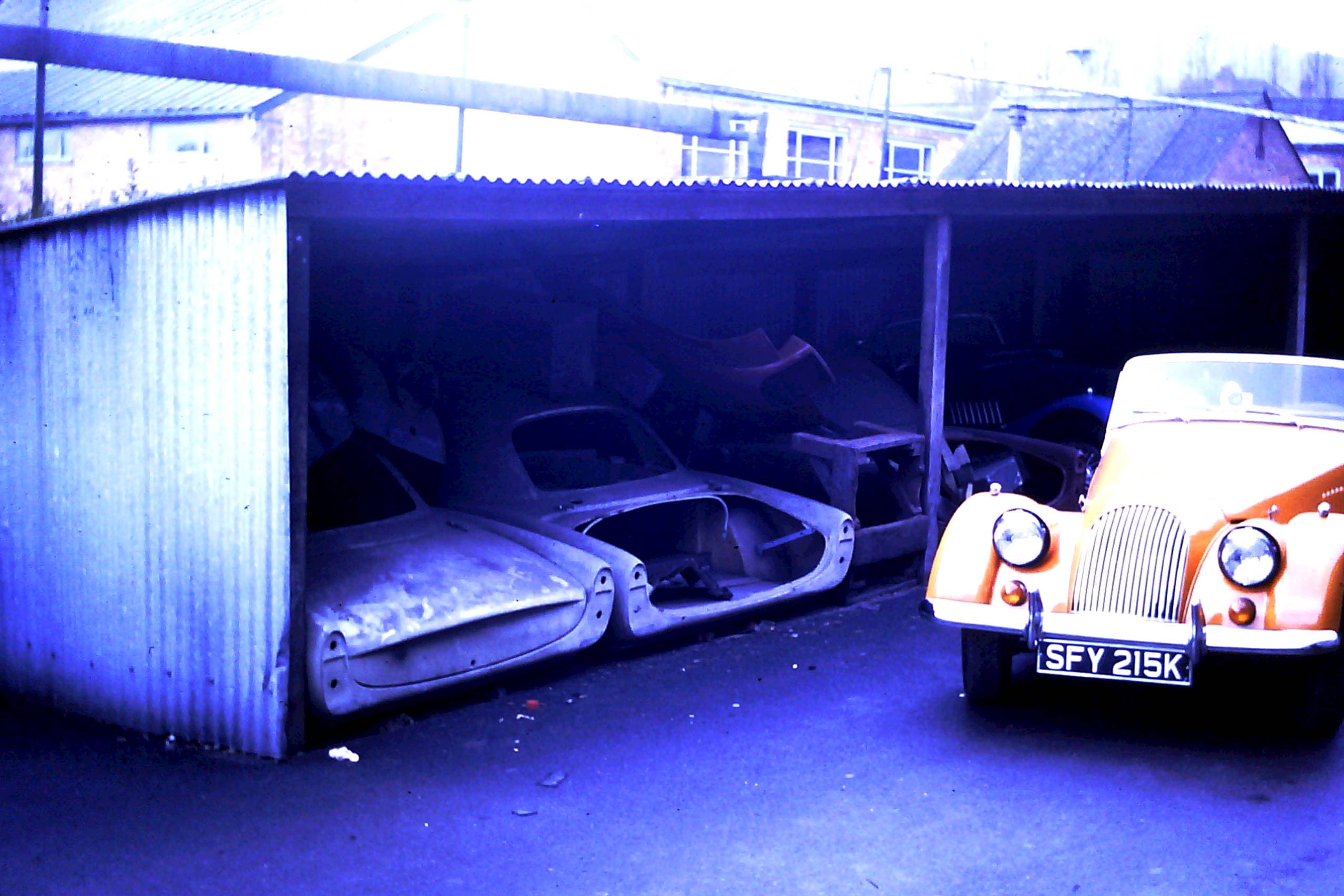 An open shed at the Morgan factory holding two unused fibreglass Plus 4 Plus shells, with an orange Morgan parked alongside